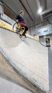 Person im Rollstuhl mit Helm und Schutzausrüstung fährt eine steile Rampe im Skatepark hinunter.