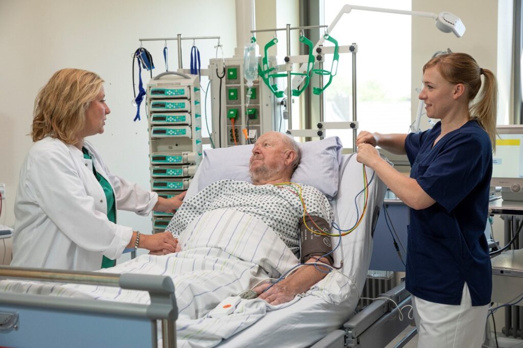 Älterer Herr im Krankenbett mit einer Ärztin und einer Krankenschwester neben dem Bett
