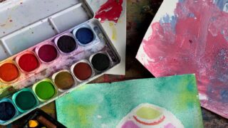 Gemaltes Bild eines Ostereis mit einem Tuschkasten und Wachsmalstiften