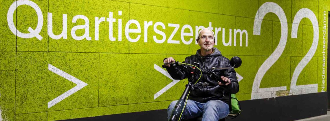 Ein Mann mit Brille und Mütze fährt auf einem elektrischen Dreirad durch das Quartierszentrum Huckelriede.