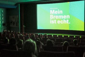 Kinoleinwand mit grün-blauem Hintergrund und dem Schriftzug „Mein Bremen ist echt.“; Zuschauerinnen und Zuschauer sitzen im dunklen Saal.