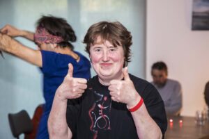 Eine Frau mit kurzem braunen Haar zeigt beide Daumen nach oben und lächelt, im Hintergrund zwei weitere Personen.