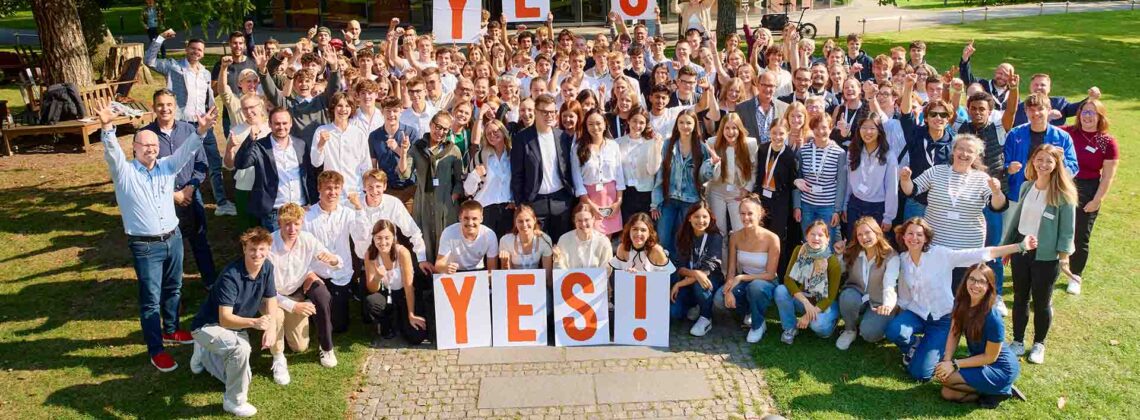 Große Gruppe junger Menschen hält YES!-Schilder und jubelt bei einer Veranstaltung im Freien.