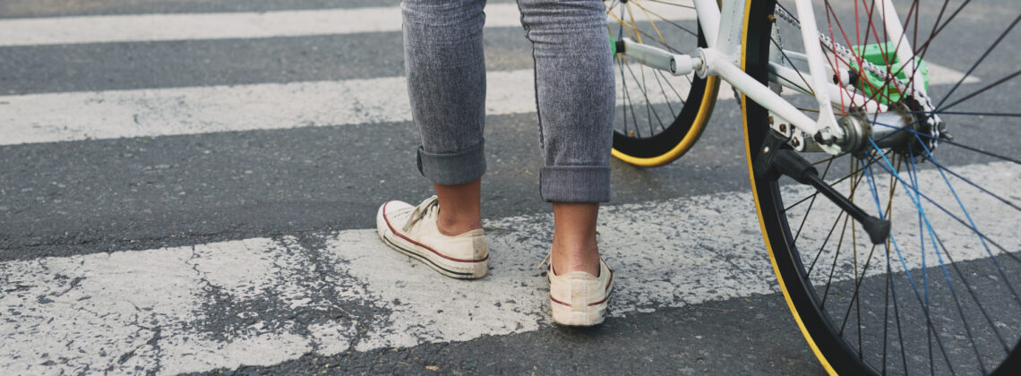 Eine Person steht mit einem Fahrrad auf einem Zebrastreifen. Nur die Beine und das Fahrrad sind zu sehen. Die Straße wirkt städtisch, Autos sind im Hintergrund zu erkennen