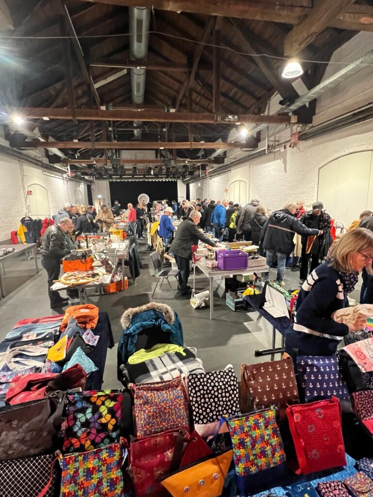 Menschen stöbern an dicht bestückten Ständen in einer Halle mit Holzbalkendecke; im Vordergrund ein Tisch mit farbenfrohen, handgenähten Taschen – aufgenommen beim maritimen Flohmarkt im Kulturbahnhof Vegesack.