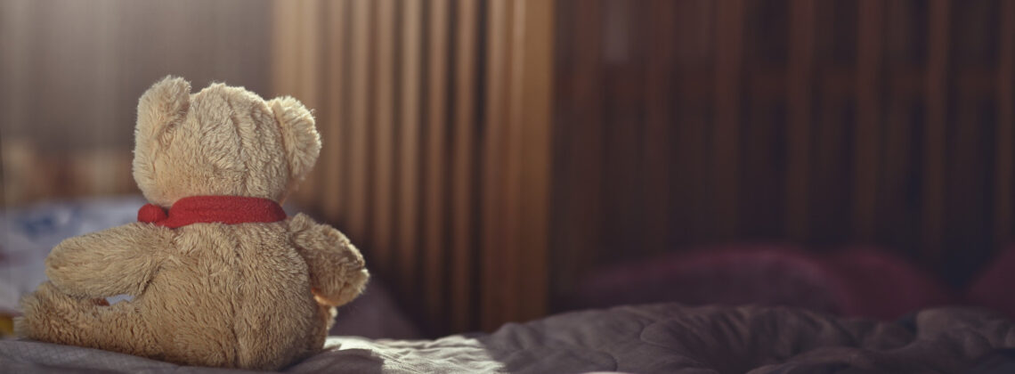Kleiner Teddybär sitzt mit dem Rücken zum Betrachter, im Hintergrund ist ein Kinderbett zu sehen - Symbolbild für Sternenkinder