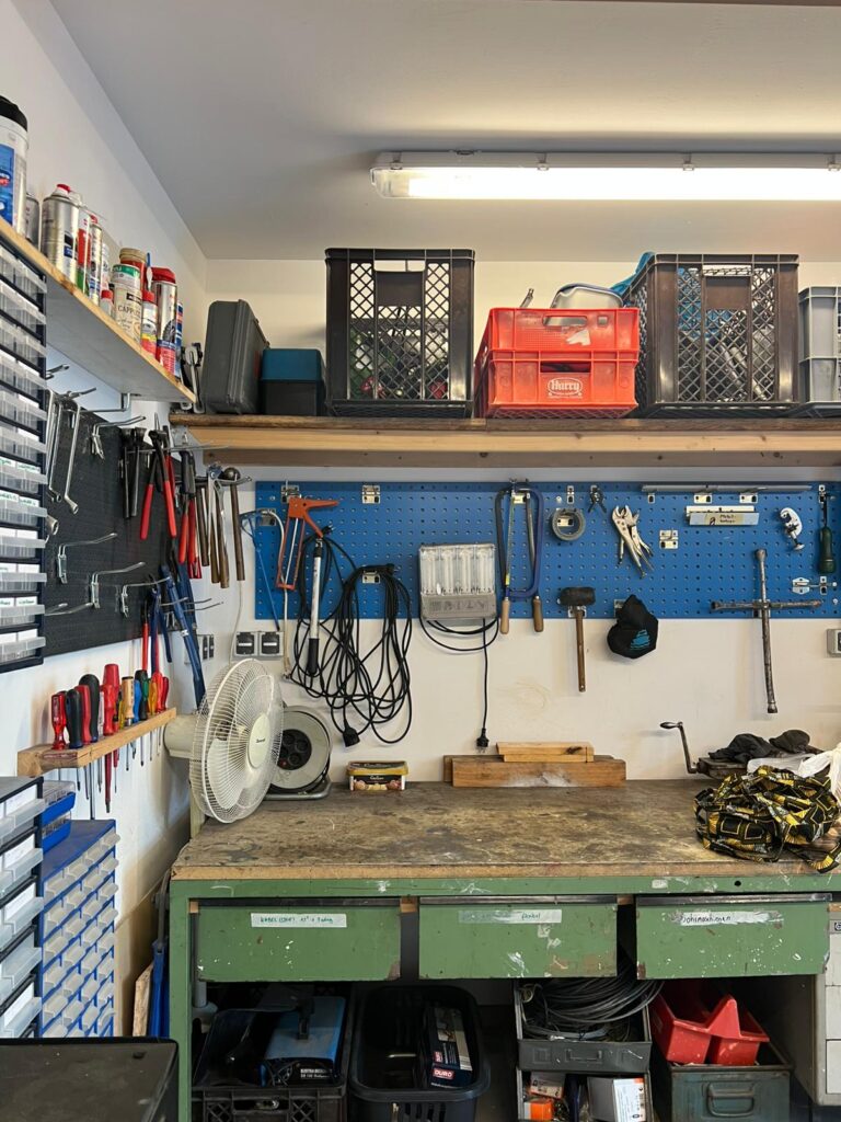 Gut ausgestattete Werkbank in einer Fahrradwerkstatt mit ordentlich sortierten Werkzeugen an einer Wand, Regalen mit Ersatzteilen und Kisten sowie einer freien Arbeitsfläche.