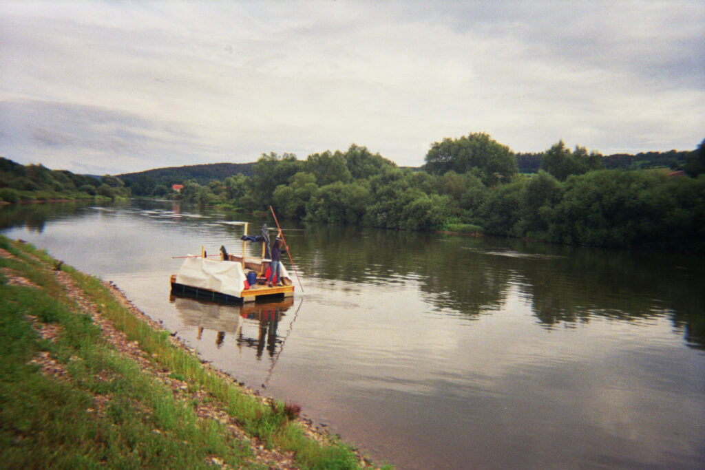 Ein Floß auf der ruhigen Weser, mit einer Person, die es mit einem langen Stab navigiert. Umgeben von grünen Ufern und sanften Hügeln.