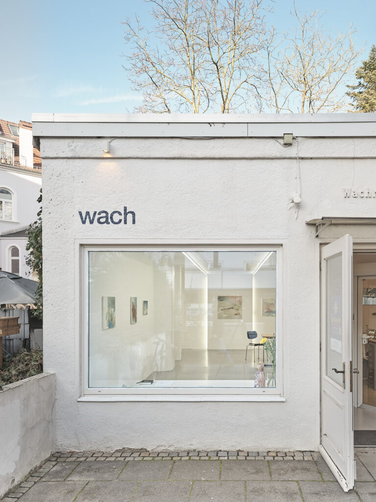 Außenansicht des Kunstraums „wach“ in der Wachmannstraße in Bremen: Ein kleines weißes Gebäude mit großem Schaufenster, durch das Kunstwerke im Ausstellungsraum sichtbar sind.