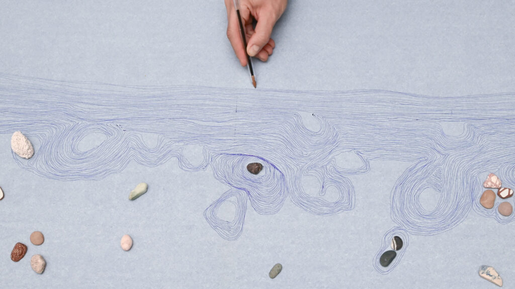 Eine Hand zeichnet mit einem Bleistift feine, geschwungene Linien auf blaues Papier; zwischen den Linien liegen kleine Steine, die Teil der Arbeit „Letter to the sea“ von Eleni Mouzourou sind.