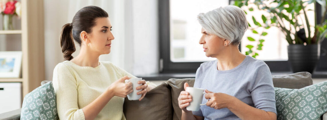Eine erwachsene Frau sitzt mit ihrer Mutter im Seniorenalter auf einem Sofa und beide halten einen Tasse in Händen