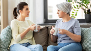 Eine erwachsene Frau sitzt mit ihrer Mutter im Seniorenalter auf einem Sofa und beide halten einen Tasse in Händen