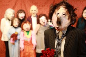 Personen mit kunstvoll gestalteten Masken und Kostümen stehen gemeinsam auf der Bühne, eine Figur hält einen Blumenstrauß