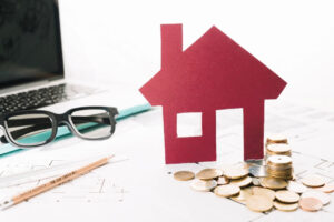 Ein rotes aus Papier ausgeschnittenes Haus steht auf einem Schreibtisch mit Laptop, Brille, Geldmünzen und Bauplänen. Symbolfoto für die Bedeutung von Eigenkapital beim Hauskauf