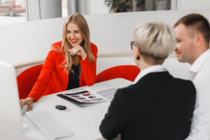 Eine Beraterin im roten Sakko lächelt und klickt mit der Maus etwas auf dem PC an. Vor ihr sitzt ein junges Paar und hört ihr zu. Symbolfoto für eine Beratung zum Immobilienkauf bei der Sparkasse Bremen