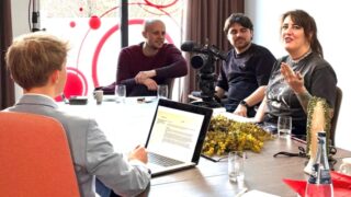 Arbeitssituation bei einem Videodreh: Vier Personen sitzen an einem Tisch, während eine Kamera auf sie gerichtet ist. Eine Person spricht und gestikuliert, während die anderen zuhören. Auf dem Tisch stehen ein Laptop, Gläser und Notizen.