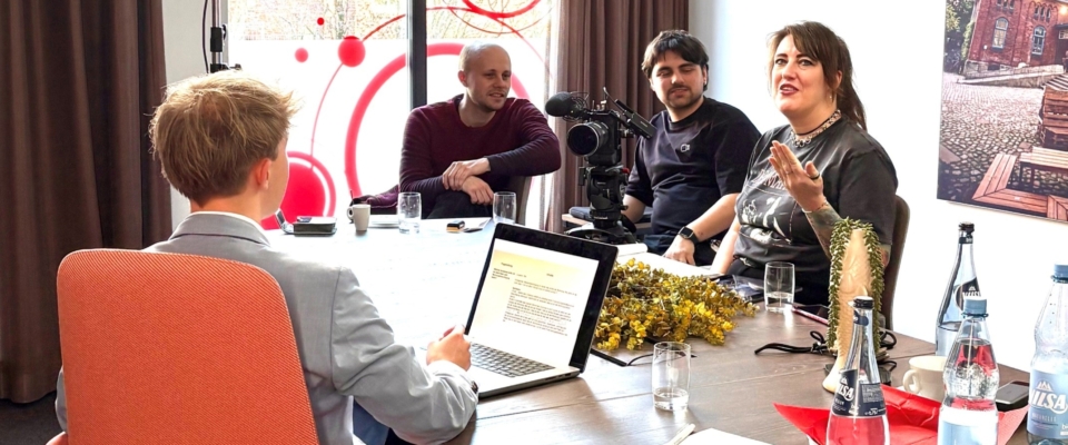 Arbeitssituation bei einem Videodreh: Vier Personen sitzen an einem Tisch, während eine Kamera auf sie gerichtet ist. Eine Person spricht und gestikuliert, während die anderen zuhören. Auf dem Tisch stehen ein Laptop, Gläser und Notizen.