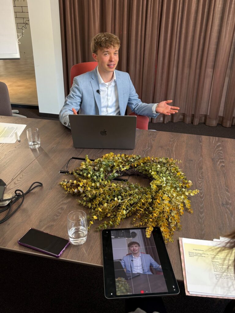 Ein Berater sitzt an einem Tisch vor einem Laptop und spricht mit offenen Gesten in die Kamera. Im Vordergrund ist ein Tablet zu sehen, auf dem die laufende Videoaufnahme angezeigt wird. Auf dem Tisch liegen zudem ein Smartphone, Unterlagen, Gläser und ein dekorativer Kranz.