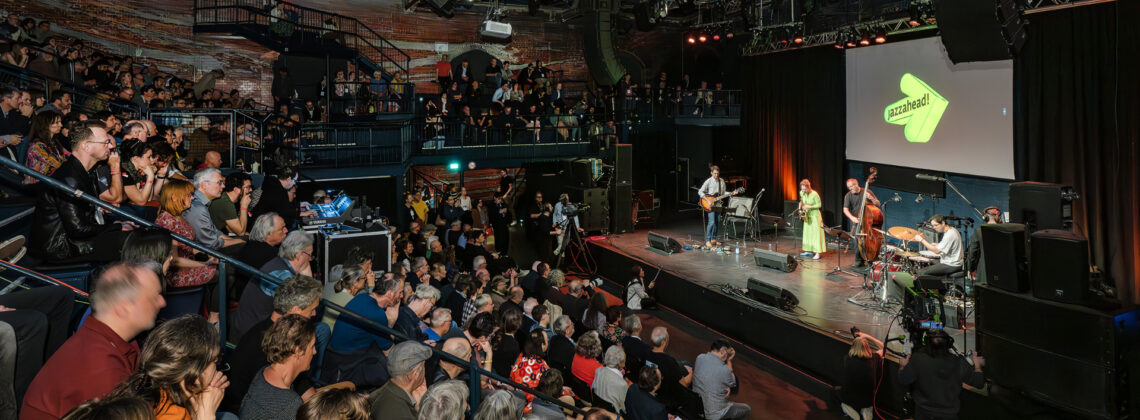 Publikum bei einem Live-Konzert während der jazzahead! in Bremen: Eine Band steht auf der Bühne, während zahlreiche Zuschauerinnen und Zuschauer das Konzert in einer voll besetzten Halle verfolgen.