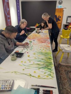 Mehrere Personen malen gemeinsam ein großes buntes Bild mit Pflanzen- und Naturmotiven auf einer langen Fläche