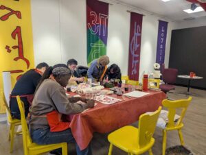 Mehrere Menschen sitzen an einem Tisch und gestalten mit Tinte und Pinseln kalligrafische Schriftarbeiten