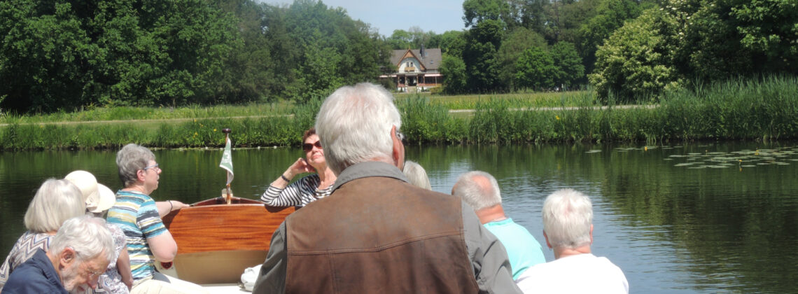 Gruppe älterer Menschen bei einer gemeinsamen Bootsfahrt auf einem ruhigen Gewässer, umgeben von grüner Uferlandschaft mit Bäumen und einem Haus im Hintergrund.