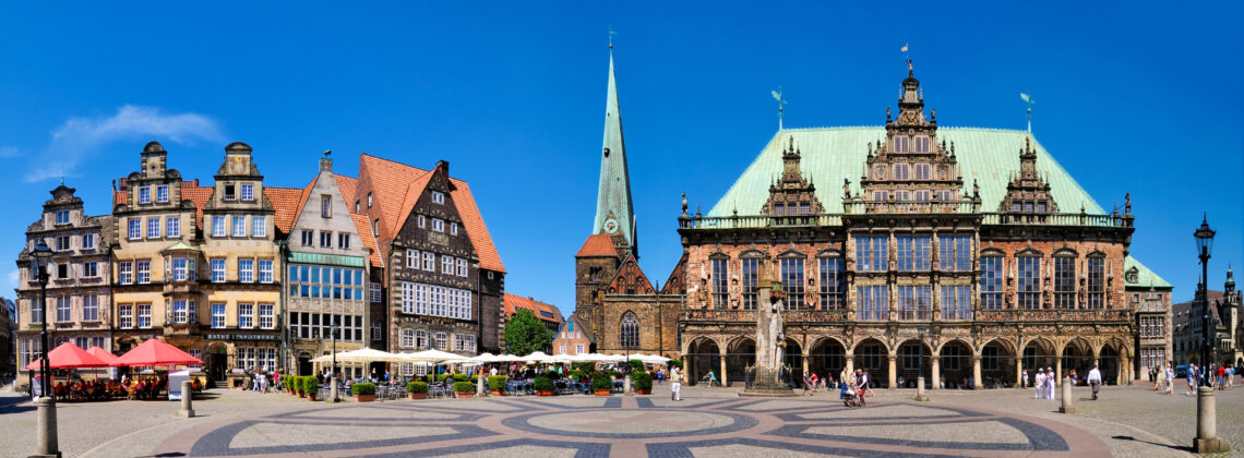Panoramablick auf den Bremer Marktplatz mit dem historischen Rathaus, der Rolandstatue und den charakteristischen Giebelhäusern bei sonnigem, blauem Himmel.