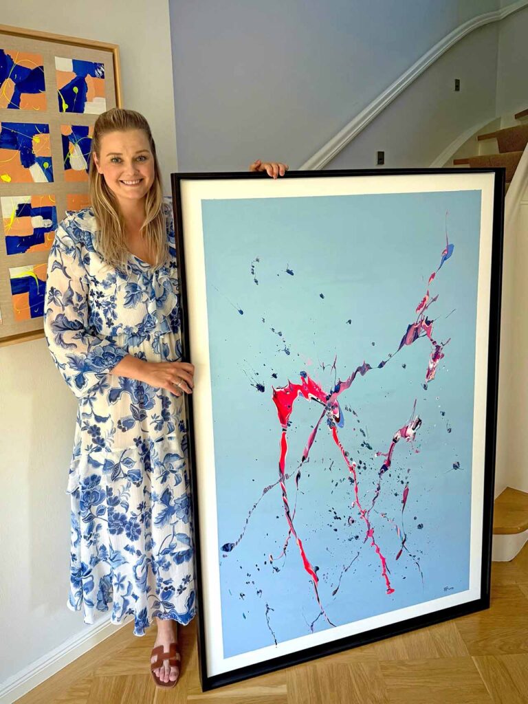 Alena Fischer steht neben einem großformatigen abstrakten Kunstwerk mit hellblauem Hintergrund und dynamischen Farbspritzern.