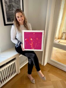Alena Fischer sitzt mit einem abstrakten Kunstwerk mit pinkem Hintergrund und orange-roten Farbbewegungen in den Händen.
