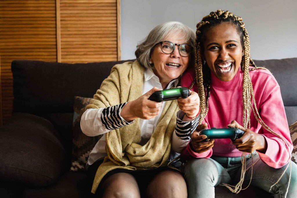 Eine ältere Frau und eine jüngere Frau sitzen zusammen auf dem Sofa und spielen gemeinsam mit Controllern.