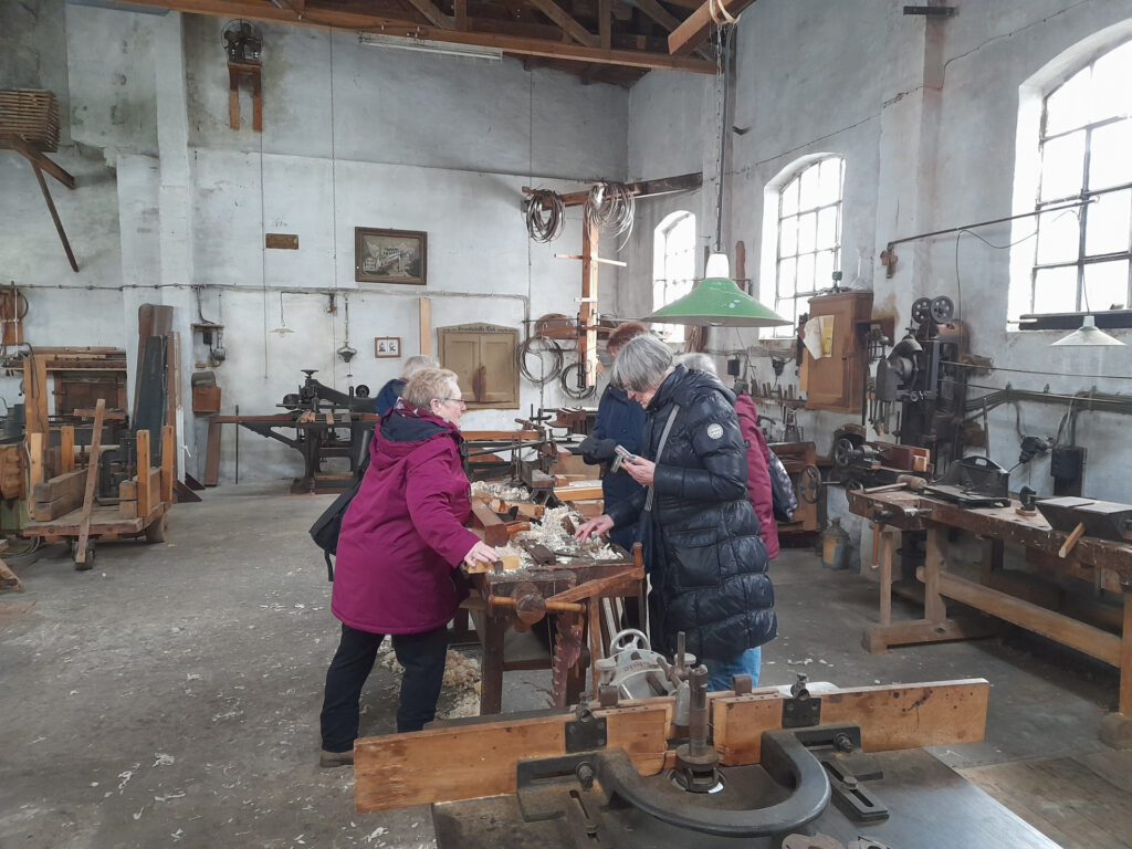 Ältere Menschen erkunden gemeinsam eine historische Werkstatt mit Holzbänken und traditionellen Werkzeugen, während sie an einem Arbeitstisch Holz bearbeiten.