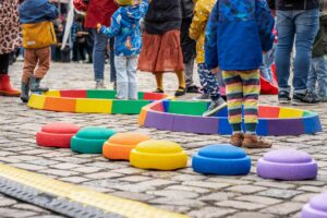 Kinder balancieren auf bunten Spielstationen auf einem gepflasterten Platz.