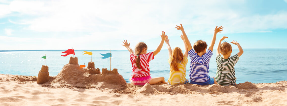 Vier Kinder sitzen am Strand neben einer Sandburg und blicken mit erhobenen Armen aufs Meer.