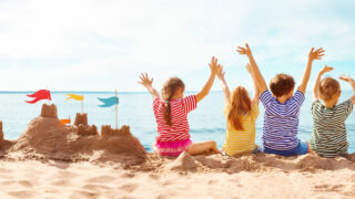 Vier Kinder sitzen am Strand neben einer Sandburg und blicken mit erhobenen Armen aufs Meer.