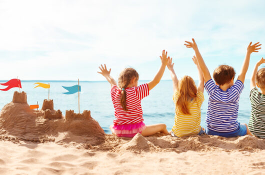 Vier Kinder sitzen am Strand neben einer Sandburg und blicken mit erhobenen Armen aufs Meer.