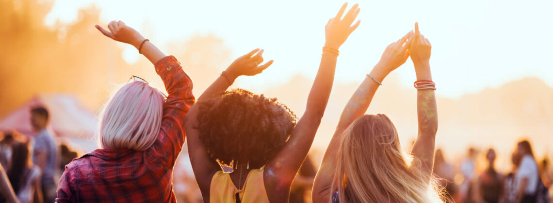 Drei junge Frauen mit dem Rücken zur Kamera, die auf einem Festival im Sommer tanzen und die Arme in die Luft strecken
