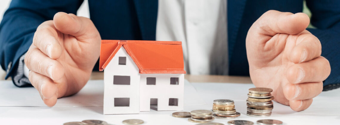 Mann im Anzug sitzt vor einem Tisch auf dem Münzen verteilt liegen und ein kleines Modellhäuschen steht, Symbolbild für Immobilien als Investition