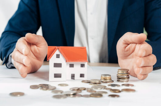 Mann im Anzug sitzt vor einem Tisch auf dem Münzen verteilt liegen und ein kleines Modellhäuschen steht, Symbolbild für Immobilien als Investition