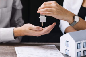 Nahaufnahme auf die Hände eines Paares, die einen Schlüssel übergeben, daneben steht ein kleines Modellhäuschen, Immobilien als Investition