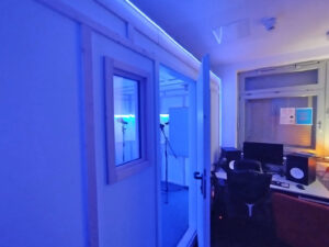 Blick in ein Tonstudio mit Schreibtisch, Lautsprechern und angrenzender Gesangskabine mit Mikrofonen.