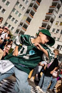 Ein Kind im Hip-Hop-Outfit nimmt am Allstyle-Dancebattle teil und tanzt vor einer Hochhauskulisse, dahinter begeistertes Publikum