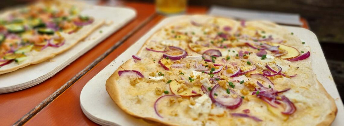 Zwei knusprige Flammkuchen auf Holzbrettern serviert auf einem Tisch im Außenbereich des Restaurants Le Feu