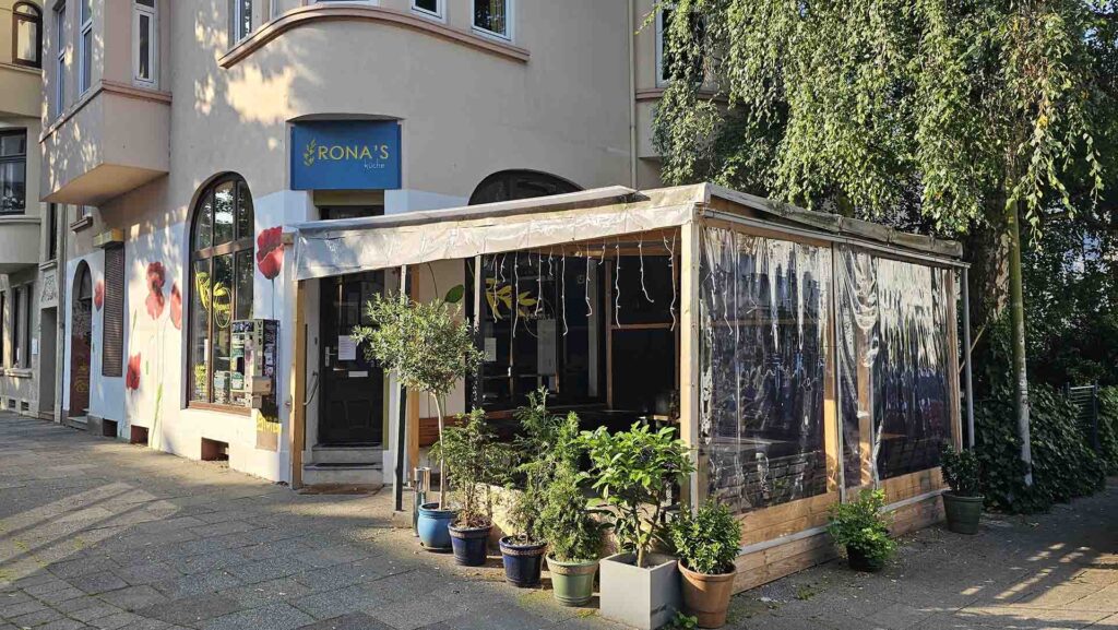 Außenansicht von Rona's Küche an einer Straßenecke mit überdachter Terrasse und Topfpflanzen im Vordergrund