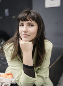 Porträt von Carla Hinrichs, die mit dem Kinn auf ihrer Hand in die Kamera blickt.