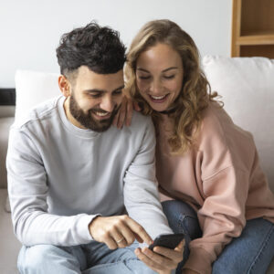 Ein Paar sitzt gemeinsam auf dem Sofa und schaut lächelnd auf ein Smartphone.