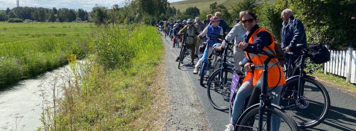 Eine größere Gruppe von Radfahrerinnen und Radfahrern steht mit ihren Fahrrädern auf einem schmalen Weg am Rand einer grünen Landschaft. Im Vordergrund lächelt eine Teilnehmerin in orangefarbener Warnweste in die Kamera, im Hintergrund zieht sich die Gruppe entlang des Weges.
