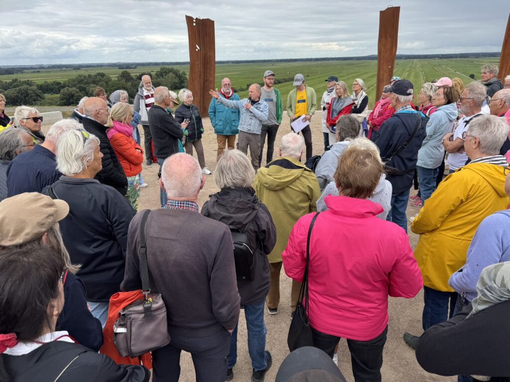Eine größere Gruppe steht am Metal Stonehenge in Bremen und hört einem Mann mit Mikrofon zu. Im Hintergrund öffnet sich der Blick über die weite Landschaft.