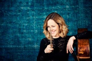 Cellistin Sol Gabetta sitzt mit ihrem Violoncello vor blauem Hintergrund und lächelt in die Kamera