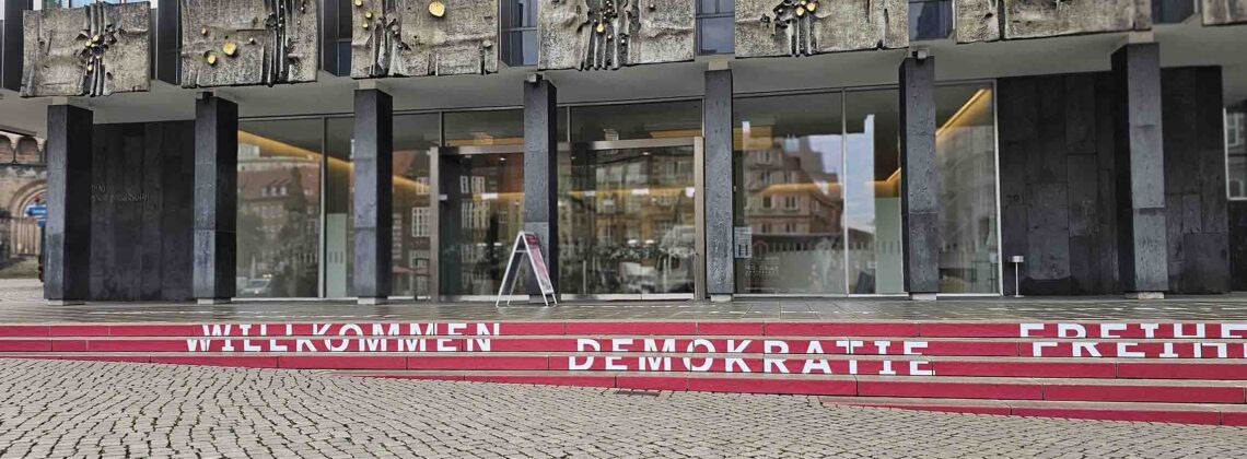 Fassade der Bremischen Bürgerschaft mit roten Stufen und den Schriftzügen „Willkommen“, „Demokratie“ und „Freiheit“.