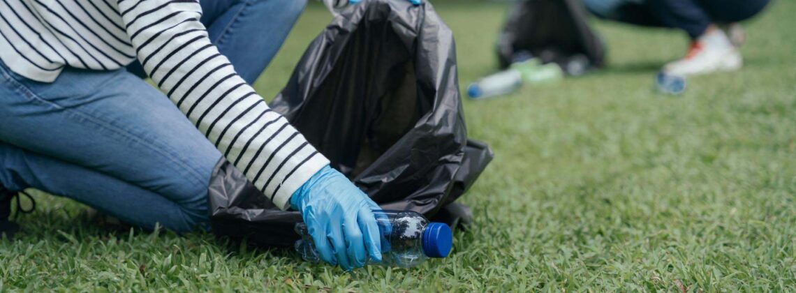 Eine Person sammelt mit Handschuhen eine Plastikflasche von einer Grünfläche auf.